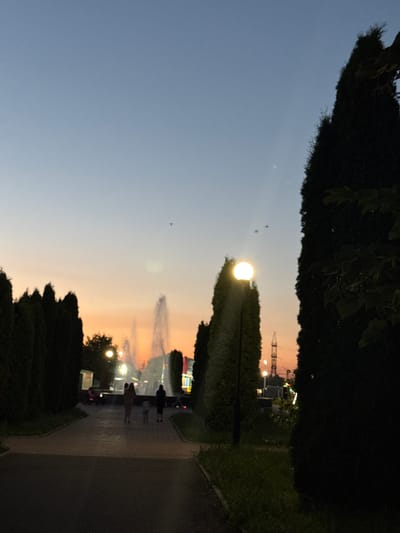 The image depicts a park scene at dusk in Endirei, Russia. A pathway runs through the park, bordered by manicured hedges and grass. Several people are visible walking along the path, their silhouettes dark against the softening sky. A bright, illuminated lamp post stands out in the foreground, casting a warm glow. In the background, a Ferris wheel and other amusement park rides are lit up, suggesting the park is a place of entertainment. The sky is a gradient of soft blue and orange, indicating the transition from day to night, and the overall atmosphere appears peaceful and serene. There is no visible text in the image.