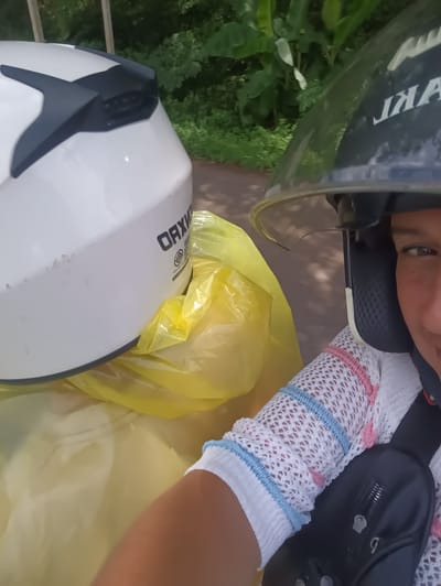 The image depicts a person wearing a black motorcycle helmet with a clear visor, positioned on the right side of the frame. They are wearing a white, open-weave sweater with light blue and pink stripes at the edges. A black over-the-shoulder bag is visible across their chest. To the left, a white motorcycle helmet with a black fin-shaped accent is contained within a yellow plastic bag. The background suggests an outdoor setting with greenery, potentially taken while moving. The text on the person's helmet, partially obscured, reads "JIA". The text on the helmet in the plastic bag reads "OAX". The location is plausibly Plai Phraya, Thailand, based on the vegetation and apparent context.