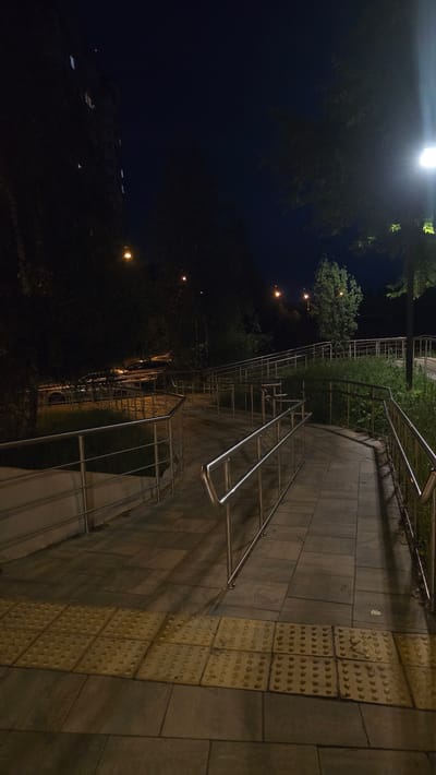 The image depicts an outdoor scene at night, featuring a paved walkway with metal railings that leads uphill and curves to the right. The walkway is constructed of large, rectangular tiles, with a section of yellow tactile paving in the foreground. Trees and green foliage line the sides of the walkway. In the background, a building with illuminated windows is visible on the left, and streetlights cast pools of light. The overall impression is one of a public access path in an urban or suburban environment. The location is identified as Razvilka, Russia. There are no people or specific actions or interactions captured in this still image. The environment is dimly lit by artificial lights.