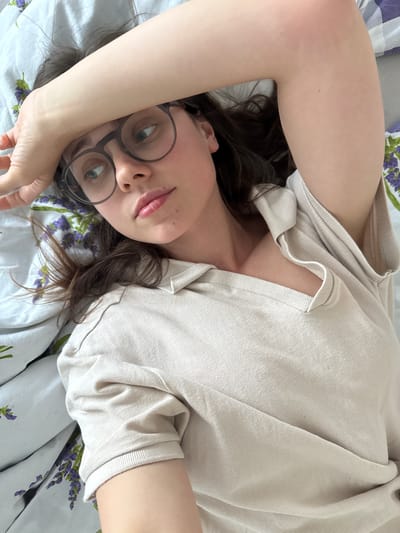 A woman with glasses is lying on a bed with a floral pattern, wearing a light-colored shirt. She has dark hair and fair skin. The location is not discernible from the image.