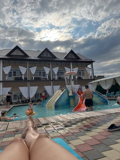 The image depicts an outdoor swimming pool area at a resort, possibly located in Yaroslavskaya, Russia, given the architectural style of the building and the presence of a water slide. The building behind the pool appears to be a multi-story hotel with balconies covered by white curtains. Several people are visible in and around the pool. A man in black shorts is standing in the water near an orange and blue water slide. A young child in a red outfit is on the slide. Another child is in the pool with an orange inflatable ring. A woman in a black swimsuit and a cap is sitting on the edge of the pool, and another person with an inflatable ring is beside her. In the foreground, a person's legs and feet are resting on a blue lounge chair, with a pair of blue clogs on the tiled ground nearby. The sky above is partly cloudy, with sunlight breaking through the clouds. The pool deck is made of multi-colored interlocking pavers.