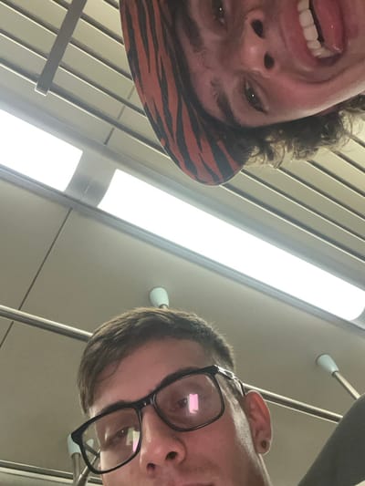 Two people are captured in a low-angle shot, with the faces close to the camera. The person on the left has curly hair, a hat with a zebra pattern, and their mouth is open, showing their teeth. The person on the right wears black-framed glasses. Overhead lighting and a ceiling with linear elements are visible in the background. The angle gives a distorted perspective, highlighting the subjects' facial features.