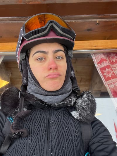 A forensic examination of the image reveals a close-up, chest-up portrait of a single individual, likely female, captured outdoors in a cold, snowy environment in Valtournenche, Italy.
The individual is wearing winter sports attire. A dark-colored ski helmet, accented with white patterns, is worn on the head, with amber-lensed ski goggles pushed up onto the helmet. A grey neck warmer or balaclava covers the lower face and neck, with visible white snow or ice particles adhering to its surface. The individual's face shows light brown eyes, dark eyebrows, and a distinct redness on the nose and cheeks, consistent with exposure to cold. The lips are pursed in a slight pout.
The torso is clad in a dark, possibly black, winter jacket featuring a textured, chevron-like quilted pattern. The jacket's hood or collar area is trimmed with dark fur, which is heavily encrusted with white snow or ice particles. Snowflakes or small ice fragments are also scattered across the surface of the jacket. Backpack straps are visible over both shoulders; the left strap features a black buckle and a small black, tubular fabric component.
The background consists of a rustic structure, likely a building. Above the individual, horizontal wooden planks suggest an overhang or ceiling. To the right, a window or glass panel reveals a red and white patterned fabric or curtain decorated with snowflake motifs. A faint, golden object is partially visible through the glass on the left side of the frame. The overall scene indicates cold, potentially snowy, weather conditions typical of a winter resort setting.