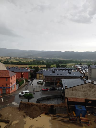 The image depicts an overcast day in Puigcerdà, Spain, with a view overlooking a town and surrounding hills. In the foreground, there is a construction site with muddy ground, piles of dirt, and construction materials including rebar and wooden planks. A blue shipping container is also present. Adjacent to the construction site, a street is wet, indicating recent rain, with a white van and a red car driving on it. Several multi-story buildings with dark tiled roofs are visible in the middle ground. One prominent building on the left is orange with multiple windows. Beyond the town, rolling hills are covered in greenery and agricultural fields. The sky is a uniform grey, with hints of lighter clouds. No people are clearly visible in the scene.