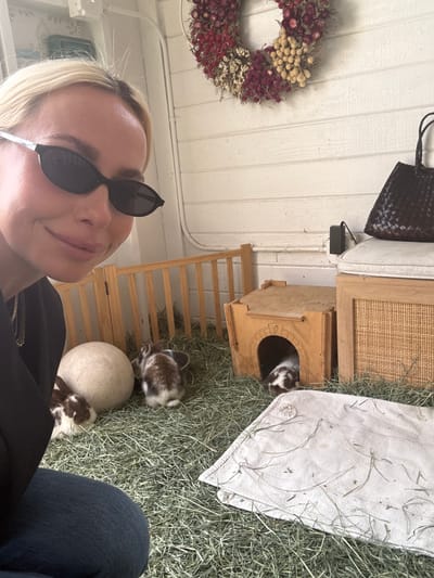 The image shows a person with blonde hair and sunglasses taking a selfie indoors with rabbits. The background includes a white-painted wall with a floral wreath, a woven black bag, a wooden cabinet, a small wooden house with a rabbit peeking out, and what appears to be a rabbit enclosure filled with hay. The environment suggests a pet-friendly or animal-related setting. There is a stone-like orb near a rabbit and a partial view of a person wearing jeans. I have no evidence of the location in the city of Solana Beach, United States of America.