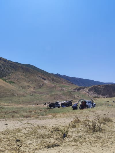 The image depicts a wide, dry, sandy expanse leading into a grassy field that stretches towards rolling, barren hills and mountains under a clear blue sky. In the middle ground, a group of people are gathered around several vehicles, suggesting a recreational outing or picnic.
Specific observations:
* **Environment:** The terrain is predominantly arid, with dry grass and scattered vegetation in the foreground and middle ground. The background features extensive, undulating hills and rugged mountains, indicative of a natural, undeveloped landscape. The sky is a uniform, cloudless blue.
* **People and Activities:** Several individuals are visible around the vehicles. Some are standing, while others appear to be seated or interacting with the cars. The presence of tents or canopies suggests an attempt at providing shade. One person is standing next to a white vehicle with their doors open. Another group is clustered around a dark-colored vehicle, with one individual holding a dark umbrella. The overall impression is of a leisurely gathering.
* **Objects:** Several vehicles are present, including a white SUV or car, a dark SUV, and possibly another dark vehicle. There are also what appear to be portable shelters or awnings set up near some of the vehicles. Miscellaneous objects, such as a blue plastic item, are scattered on the sandy ground in the foreground.
* **Location Context:** The image is identified as being in the city of Sultanabad, Tajikistan. This suggests the scene is situated within or on the outskirts of this urban area, within the context of Tajikistan's natural geography.