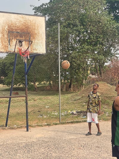 The image depicts an outdoor basketball court in Umunekwu, Nigeria. In the foreground, an asphalt court surface transitions to a chain-link fence and a grassy, unkempt area with scattered litter. Behind this, dense green foliage and trees are visible, with some drier, brown vegetation on the right.
A basketball hoop with a heavily rusted white backboard, a blue metal support frame, and a tattered red and white net stands on the left. To its right, an orange, worn basketball is suspended mid-air.
Two individuals are present. A young Black male, of lean build, stands centrally, facing slightly right and looking upwards towards the basketball. He wears a short-sleeved camouflage-patterned shirt (greens, yellows, browns), white basketball shorts with the number "14" in red on the right leg, and dark slide sandals. A second individual, partially visible on the far right, is a Black male wearing a black and green sleeveless jersey; only his right shoulder, arm, and part of his back are within the frame. A thin, grey metal pole stands vertically behind the chain-link fence, to the right of the basketball hoop.
