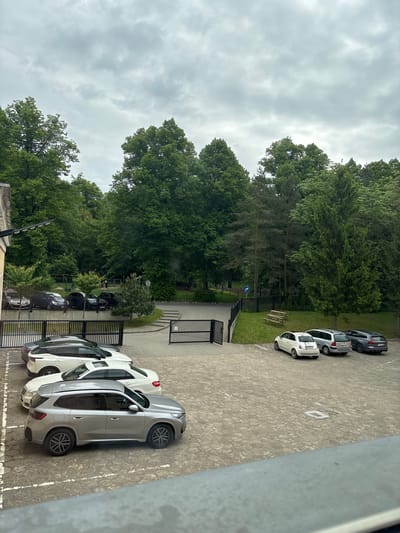 The image shows an outdoor scene with a parking lot in Gryfino, Poland. The parking lot is partially filled with several cars of varying colors, including white, silver, and dark grey. A metal fence separates the parking lot from a green area with trees and shrubs. In the background, there is a playground with play equipment. The sky is overcast and grey. The image appears to be taken from an elevated position.