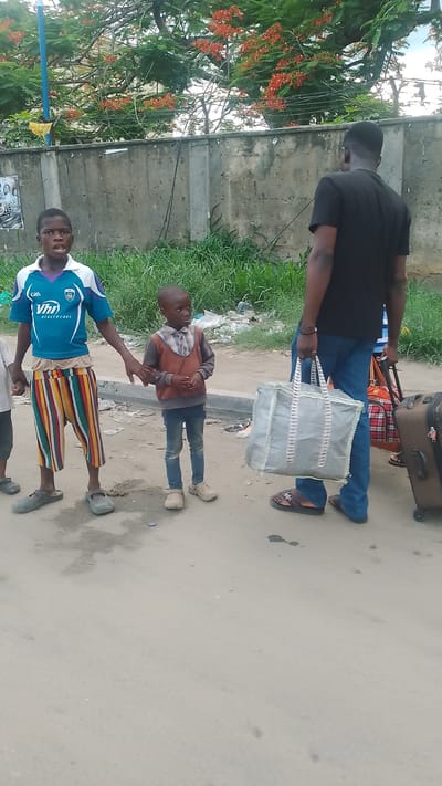 In Effurun, Nigeria, the image depicts a group of people standing on a paved road. Two children are holding hands near the left side of the frame, while an adult male carries a large bag and pulls luggage on the right. Trash and vegetation are visible along a concrete wall in the background. A tree with orange flowers is visible in the upper portion of the image.