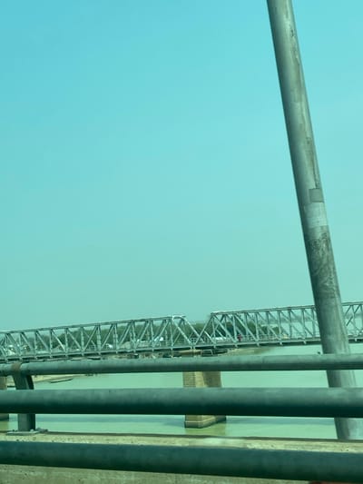 A structure, possibly a bridge, is visible through vertical pipes. The bridge has a latticed design. The sky is a muted blue and contrasts with the greenish water underneath the bridge. Additional metallic pipes run horizontally across the top of the frame, obscuring the view of the structure.