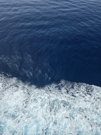 The image shows a close-up view of the sea with the wake of a boat. The water is a deep blue and the wake is white and frothy, with small bubbles and ripples. The sunlight reflects off the surface of the water, creating a shimmering effect. There are no people or man-made objects visible in the image. The scene is likely captured from a moving vessel, suggesting a maritime environment. The location of Santa Ponça, Spain, is a coastal town known for its marina and beaches, making this a plausible setting for such a view.