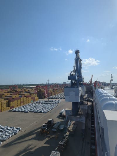 This is a high-angle, vertically oriented photograph taken at the Port of Hambantota in Sri Lanka. The scene depicts a busy port terminal under a clear blue sky with scattered white clouds, suggesting a bright, sunny day.
The primary subjects are large shipping containers stacked high and rows of white vehicles parked on the tarmac. Several industrial cranes are visible, including a large blue gantry crane in the foreground and red cranes further in the background, indicating active cargo handling operations. The presence of the ships, cranes, containers, and vehicles strongly suggests the import or export of goods and automobiles.
Notable details include the text "ZPMC 40t-40m" on some of the crane structures, which likely refers to the manufacturer and capacity of the equipment. The parking area for the vehicles is marked with painted lines and numbers (e.g., "061", "062", "070", "071", "072", "090", "1000"), suggesting an organized system for vehicle storage. The edge of a large white ship, possibly a car carrier, is visible along the top right of the image. The overall impression is one of a functional and large-scale logistics hub.