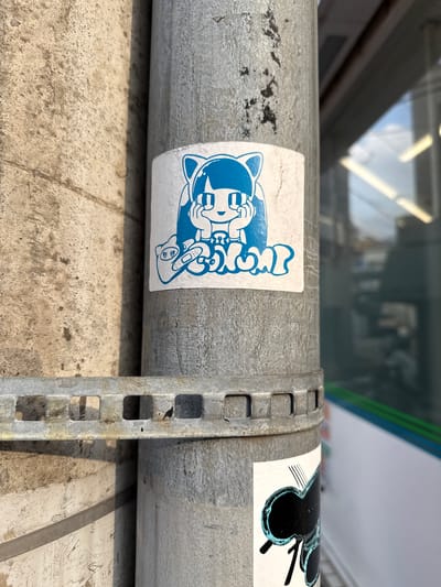 A blue and white sticker featuring a cartoon cat-like character with large eyes and cat ears is affixed to a silver metal pole. The character is depicted with its hands near its face and is wearing a small bandage on its left paw. Below the character, text is visible in a bubbly font. To the left of the pole, a textured concrete surface is present, and a metal bracket with square openings is attached to the pole. In the background, a blurred reflection of a building with horizontal windows and lights is visible, suggesting an urban setting, likely a street or sidewalk in Toshima, Japan. Another sticker, black and teal, is partially visible below the cat sticker.