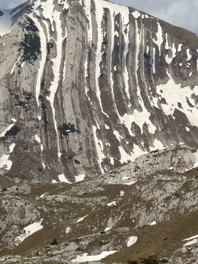 The image depicts a mountain landscape in Boričje, Montenegro. The left side of the image features a layered rock formation, with bands of gray stone interspersed with patches of white snow. The right side of the image shows a more rugged, uneven terrain, with a mix of gray rocks, green vegetation, and scattered patches of snow. There are no people visible in the scene. The environment appears to be a high-altitude, rocky area, typical of mountainous regions.