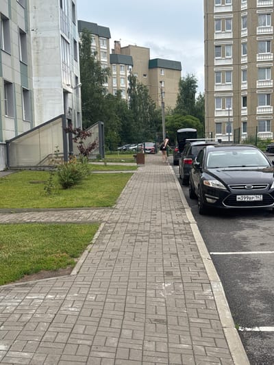 The image depicts a view of an apartment building in Bugry, Russia, with a paved walkway leading towards it. Several parked cars are visible along the walkway, with the trunk of one car open. A person, likely a woman, is standing near the open trunk, possibly interacting with it. The environment appears to be an urban setting, with buildings and greenery surrounding the walkway and parking area. The sky is overcast.