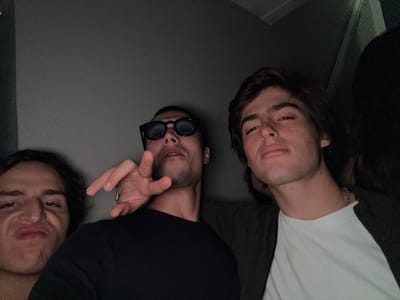 The image displays a close-up, dimly lit scene featuring three young adult males in an interior setting. No specific location within South Bend, United States, is discernible from the available visual information.
From left to right:
1. A male subject, with only the left side of his face and dark, wavy hair visible, making a "duck face" or pouting expression.
2. A central male subject, wearing dark sunglasses and a black t-shirt. He has short facial hair and is looking towards the camera with his mouth slightly open. His right hand is raised, fingers splayed and slightly bent, pointing in the general direction of the camera. A silver-colored ring is visible on his left pinky finger.
3. A male subject positioned on the right, wearing a white t-shirt and a dark outer garment. He has dark, medium-length hair and a neutral expression, looking directly at the camera. A gold-colored chain is visible around his neck.
The background consists of a uniform dark gray surface, indicative of a wall or similar structure in a poorly lit room. The overall lighting is low, casting soft shadows and limiting detail. The composition suggests a close-range photograph, potentially a selfie.
