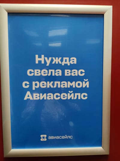 The media file is a photograph of a framed blue poster with white text. The poster is hanging on a red wall. The text on the poster reads: "Нужда свела вас с рекламой Авиасейлс". Below this text, there is a logo and the words "авиасейлс".

The poster is centrally framed within the image. The white border of the frame is visible, and the red wall serves as the background. The lighting in the image is somewhat diffused, creating subtle reflections on the poster's surface. The overall impression is that of an advertisement displayed in a public space, possibly an airport or a travel agency, given the text "Авиасейлс" which likely refers to an airline or a travel booking service.

The text "Нужда свела вас с рекламой Авиасейлс" translates to "Need brought you to Aviasales advertising." This suggests the poster is aimed at people who are looking for travel services, implying that Aviasales can meet their needs. The logo, though small, appears to be a stylized camera lens or aperture, which might be related to capturing memories or experiences associated with travel. The location context of Khrabrovo, Russia, suggests this might be a poster displayed in Khrabrovo Airport or a related facility in that region. There are no people visible in the image, and the focus is entirely on the poster and its message. The overall mood is informative and promotional.