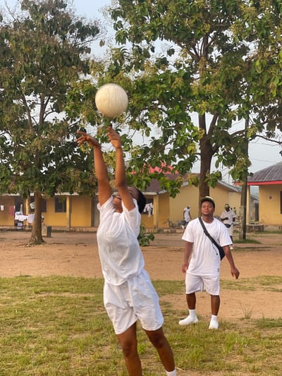 In Emure-Ekiti, Nigeria, two individuals are engaged in a volleyball game outdoors. The foreground features a person in mid-air, reaching to hit a volleyball above their head. This person is wearing a white t-shirt and white shorts. Behind them, another person stands watching, also in a white t-shirt and white shorts, with a dark bag slung across their chest. The background reveals yellow buildings with red roofs, characteristic of local architecture. Lush green trees with visible foliage and small seed pods dot the landscape, suggesting a tropical or subtropical environment. The ground is a mixture of dirt and sparse green grass. Several other individuals in similar white attire are visible in the distance, some wearing green caps, possibly part of a group or organization. Clothes are hanging on a line outside one of the buildings. The sky is overcast, indicating diffused lighting.