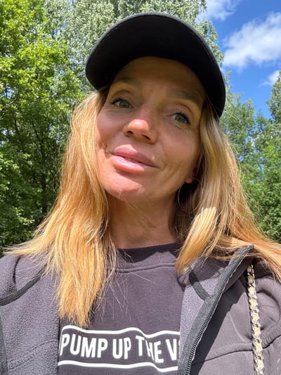 A medium shot photograph shows a woman wearing a black baseball cap and a dark gray zip-up hoodie over a black sweatshirt with white text. The woman has blonde hair, fair skin, and is looking directly at the camera with a slight smile. She is wearing large hoop earrings. The background is a bright, sunny day with green trees and a blue sky with white clouds. The photograph was taken outdoors, likely in a park or natural area. There is no indication of the specific location within the city of Saint Petersburg, Russia.