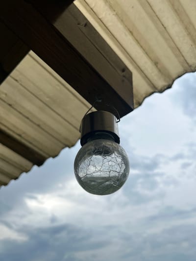 The image shows a close-up, low-angle shot of a cracked glass light bulb mounted to a structure. The light bulb is round and appears to be made of clear glass with a network of fine cracks across its surface. It is attached to a metallic fixture, which is further connected to a wire.
The structure the light bulb is attached to is made of corrugated, translucent material, likely plastic or fiberglass, supported by wooden beams. The corrugated material is light brown and weathered, showing some discoloration and dirt. The wooden beams are dark brown and appear to be rough-hewn. The perspective is looking upwards, with the sky filling the background.
The sky is overcast with diffused sunlight filtering through the clouds. The clouds are a mix of grey and white, suggesting a day with variable weather. The lighting is soft, indicating it is likely daytime, possibly late morning or afternoon.
There are no people or animals visible in the image. The focus is on the light bulb and the surrounding structure, with the sky as a backdrop. The overall mood is somewhat stark and industrial, with the cracked bulb perhaps hinting at a state of disrepair or age. The image was captured in Haaslava, Estonia. There is no visible text in the image.
