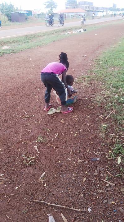 The scene is captured at a roadside location within Siaya Township ward, Kenya. In the immediate foreground, an individual with dark skin and dark braided hair, clad in a pink t-shirt and dark trousers, is observed bending over. They are barefoot, with one pair of pink flip-flops and one pair of light green clogs positioned on the reddish-brown, gravelly ground near their feet. This individual is attending to a younger child, also with dark skin, who is seated inside a multi-colored cardboard box. The ground is sparsely covered with dry leaves, small sticks, and various small pieces of litter.
Further into the midground, a paved road stretches horizontally. Multiple individuals are present along the road and its adjacent dirt shoulder. Two individuals are riding a motorcycle, and another is riding a bicycle. Several other indistinct figures are visible walking in the distance. The environment includes patches of green grass along the roadside, various small, simple structures, and distant trees. In the far background, a prominent multi-story concrete building is visible under an overcast sky.