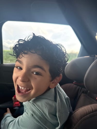 A young male subject, approximately primary school age, with dark, curly hair and a light complexion, is depicted in the rear passenger compartment of a vehicle in Pourrières, France. He is smiling broadly, mouth open, revealing a full set of primary teeth, and looking directly at the camera. The subject is wearing a light blue hooded sweatshirt. Behind him, brown upholstered vehicle headrests are visible. Through the vehicle's window, an exterior scene shows an overcast sky and a distant green landscape.