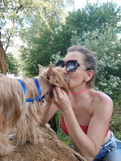 A woman with her hair pulled up and large sunglasses is leaning over a fallen log, kissing a small, tan-colored dog. The dog is wearing a blue and red harness. The woman is wearing a red strapless top and blue jeans. The background is filled with lush green trees and foliage. The scene appears to be outdoors in a wooded area, possibly a park or natural reserve. The location is not specified as Fyodorovskaya, Russia, nor is there any information to suggest this.