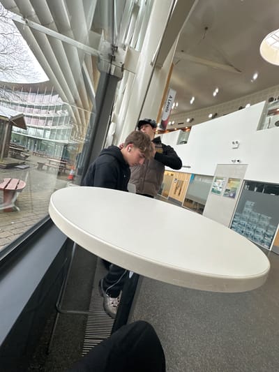 The image shows an indoor scene in what appears to be a student area, likely within a school or college setting, potentially within the city of Wistaston, United Kingdom. Two young individuals are partially visible at a round white table. One is wearing a black jacket and appears to have an earbud in their ear. The second person is partially obscured, wearing a dark top with a gold logo and a brown puffer jacket. A sign stating "A Levels" is visible in the background. The scene is observed through a window overlooking an outdoor seating area with wooden benches. The location appears to be modern with high ceilings and large windows.