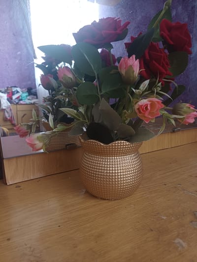 The image shows a bouquet of artificial red and pink roses in a textured golden vase. The vase sits on a wooden surface, likely a table or dresser. Behind the vase, a mirror reflects the bouquet, and in the reflection, a blurry background shows what appears to be a wooden dresser with clothing draped over it. The overall setting suggests an indoor space. There is no information available that can locate this scene in Jumashuy, Uzbekistan.