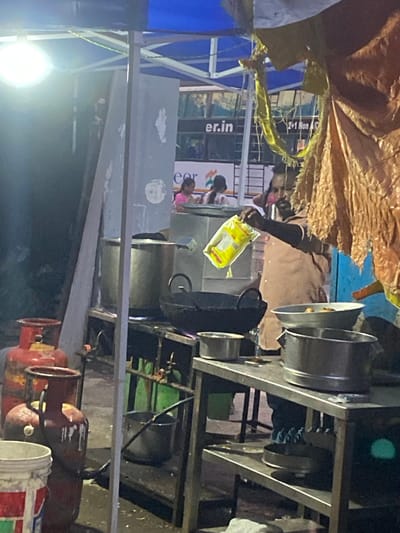 The image shows an outdoor scene, possibly a street food stall or a small eatery, in St. Thomas Mount-Pallavaram Cantonment Board, India. The lighting suggests it is either dusk or nighttime, with artificial lights illuminating the area.

The main subjects are people and objects related to food preparation and serving. In the foreground, there are several large, red gas cylinders, indicating an operational cooking setup. Above them, a metal structure supports large cooking pots or containers, with at least two visible. A person, partially visible and wearing a light-colored shirt and dark sleeves, is holding a yellow packet of what appears to be food seasoning or instant noodles. This suggests food preparation is actively underway.

In the background, there are glimpses of a structure with blue and yellow tarpaulins, possibly a canopy or part of the stall's roofing. There are also hints of other people, including children, in the background, adding to the sense of a public or commercial setting. One child is seen in a pink top, and another in a dark top.

Visible text includes "er.in" on a white banner or sign in the background, and part of an Indian flag emblem is also visible. The overall atmosphere seems to be one of a busy, potentially crowded, outdoor food vendor environment. The image is taken from a slightly elevated angle, looking down and across the scene.