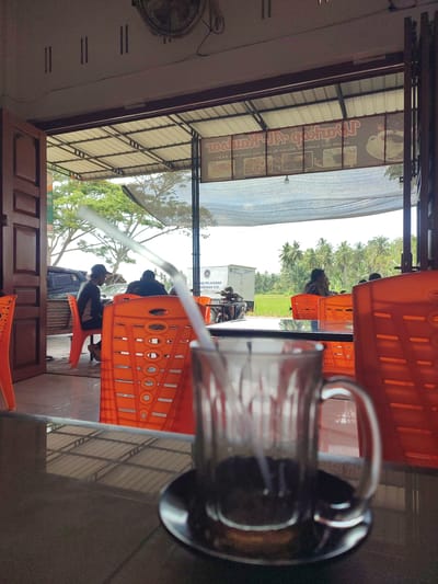 This image depicts a casual, open-air eatery or cafe in Kruengmanyang, Indonesia, observed from an indoor perspective. In the blurry foreground, a glass mug containing a dark liquid and a white straw rests on a dark table.

The midground features several vibrant orange plastic chairs, some occupied by patrons. People are seen relaxing in these chairs, facing outwards towards the bright exterior. One person on the left, wearing a dark shirt and cap, is partially visible, while another with a patterned shirt sits further back.

The background reveals a bright, sunny daytime scene with a lush tropical landscape. Numerous green trees, including a prominent cluster of palm trees, line the horizon. A green field, possibly a rice paddy, is visible before the distant trees. A white truck, bearing the partially visible text "AN PELAYANAN," "MENUHAN GIZI," and "NASIONAL," suggests a public service vehicle. A motorcycle is also partially visible near the truck.

The establishment has an open design with a light-colored corrugated metal roof supported by a dark frame, and a concrete floor. A ceiling fan is partially visible in the top left. Mirrored text from an overhead banner, reading "zostwa-JPqötu" and "1810930ОХ АЯА МАВ HOAX TOJ KAJAL," is reflected on the table surface. The clear, bright sky indicates a sunny day, consistent with a warm climate. The overall mood appears relaxed.