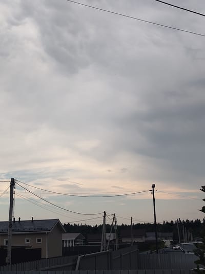 The image depicts a cloudy sky over a rural or suburban area with several houses and utility poles. The houses have pitched roofs, and some appear to be under construction or recently built. Fences are visible in the foreground, and a dense line of trees forms the background. Overhead power lines crisscross the frame. The overall lighting suggests it is daytime, but the heavy cloud cover creates a subdued, overcast atmosphere. The location is identified as Skurygino, Russia.