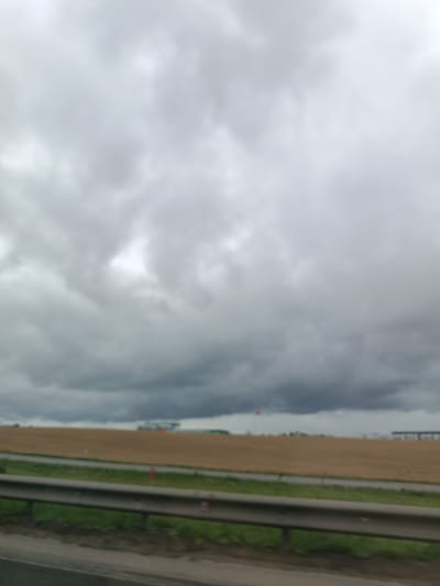This is a blurry, low-resolution photograph taken from a vehicle, likely on a highway or major road. The scene depicts a rural landscape under a cloudy, overcast sky, suggesting a cool or damp day.
In the foreground, a metal guardrail runs horizontally, indicating the viewer is situated next to a road. Beyond the guardrail, a strip of green grass is visible, followed by a wide expanse of dry, brown earth, possibly a harvested field or agricultural land. A smaller green strip runs parallel to the brown field, suggesting a road or path. In the distance, there are hints of structures, including what appear to be industrial buildings or storage facilities, some with visible roofs and supports. There is also a tall, thin structure that might be a tower or chimney, and a red flag is visible on a pole.
The sky is dominated by thick, dark grey clouds, with some lighter patches suggesting the sun is behind them, but not breaking through. This creates a somber and somewhat dramatic atmosphere. The time of day is indeterminate due to the heavy cloud cover, but it appears to be daytime. The location cues, such as the rural landscape and the type of buildings in the distance, combined with the provided location context of Путрышкаўскі сельскі Савет, Belarus, suggest an agricultural or industrial area in the Belarusian countryside. No people are discernible in the image, nor is there any visible text. The overall impression is one of a vast, somewhat desolate, and weather-beaten landscape.