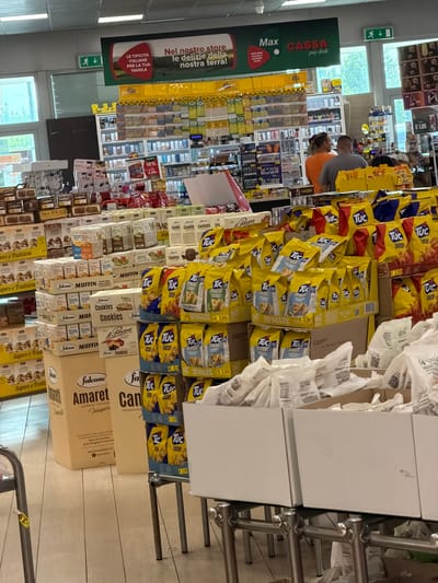 An interior shot of a supermarket displays shelves stocked with various food products, predominantly baked goods and snacks. In the foreground, large boxes of cookies and amaretti biscuits are visible, with "Amaretti Morbidi Italiani" and "Cane di Zucchero" branding on the packaging. To the right, stacks of yellow and blue bags labeled "Tuc Crisp" and "Tuc Sfoglie Naturale" are arranged on shelving units, with prices like "€ 2.99" displayed. In the background, shelves are filled with packaged goods, including what appears to be tobacco products, with signage indicating "Max CASSA pay desk". Two individuals, a man in an orange shirt and another in a grey t-shirt, are seen standing near the checkout area. The overall environment suggests a retail setting within a supermarket in Due Carrare, Italy, given the presence of Italian product labeling and the "CASSA" (cashier) signage common in Italian stores.
