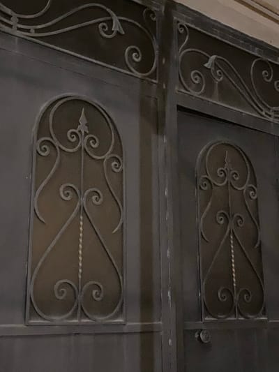 The image shows a close-up view of an ornate metal gate. The gate is dark gray and features decorative scrollwork. There are two rectangular panels, each containing an oval-shaped cutout with intricate metal detailing. Within these cutouts, curved metal lines form swirling patterns, resembling leaves or tendrils. A twisted metal rod runs horizontally across the center of each oval. To the left of the main panels, a vertical section of the gate displays a similar decorative motif, with curved lines and small circular accents. The metal appears to be wrought iron or a similar material, with a matte finish. The lighting suggests it is either indoors or outdoors in ambient light, as there are no strong shadows. The overall impression is one of craftsmanship and decorative design. The location context is Verin Artashat, Armenia, which could imply the style of the gate is typical of the region, though this is not explicitly visible in the image itself. No people or text are visible.