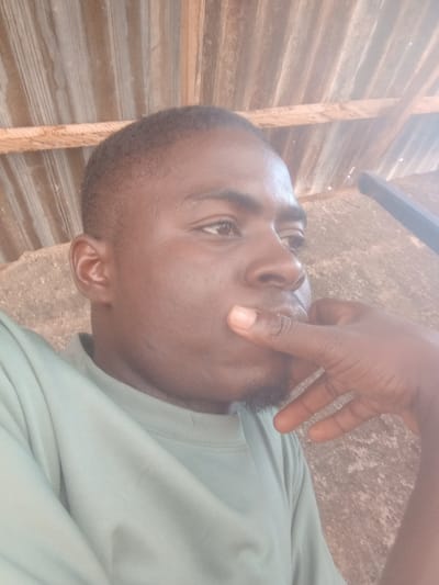 A medium close-up shows a Black man, appearing thoughtful, with his right hand placed on his chin. He has short, dark hair and is wearing a light green t-shirt. The background includes a corrugated metal roof and what appears to be a concrete wall. The photo seems to have been taken outdoors. Specific details pinpointing the exact location within Umuokpu, Nigeria, are not discernible from the image.