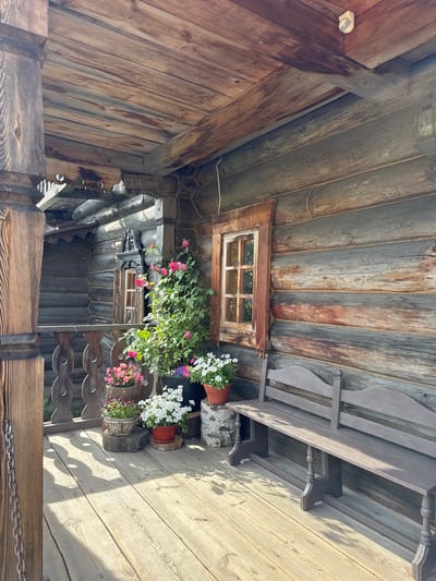 The image shows an outdoor scene with wooden structures and potted flowers, suggesting a rustic or garden setting. The perspective is looking upwards, likely from a seated or low position. The primary subjects are the weathered wooden walls and ceiling, a small window with multiple panes, and several potted plants with blooming pink and white flowers. There's also a wooden bench or railing visible, with decorative elements. The time of day appears to be sunny, indicated by the strong shadows cast on the wooden floorboards on the right side of the image. The overall atmosphere is calm and serene. There are no people visible in the image. The location context of Nevskii Parkleskhoz, Russia, suggests this might be part of a park, a dacha, or a similar outdoor recreational area.