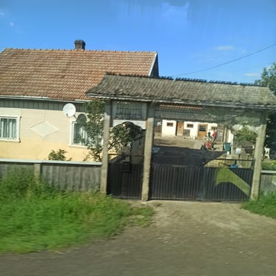 The image shows a rural property with a house and a gate, located in the city of Grănicești, Romania. The house is a single-story building with a tiled roof and pale yellow siding. It has two visible windows with white frames and a white satellite dish mounted on the exterior wall. The property is enclosed by a wooden fence and a large metal gate. Beyond the house, a smaller structure with a weathered roof is visible, and further in the background, a few more buildings and trees can be seen. The sky is clear and blue. The overall impression is that of a modest, well-maintained rural residence.