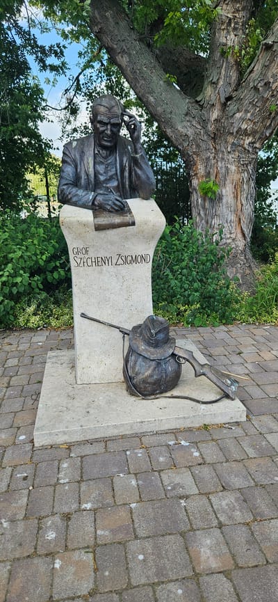 The image shows a bronze statue of a man seated with his hand on his head, holding a pen, above a stone pedestal inscribed with "GROF SZÉCHENYI ZSIGMOND". In front of the pedestal, on the ground, are bronze sculptures of a hat, a bag, and a rifle. The statue is located outdoors, with a large tree behind it and green foliage surrounding the base. The ground is paved with interlocking bricks. The overall scene appears to be a monument dedicated to Graf Széchenyi Zsigmond in the town of Balatongyörök, Hungary.