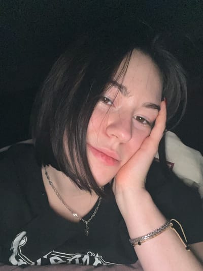 A woman with short, dark hair rests her face on her hand, appearing relaxed. She wears a dark t-shirt and a delicate silver necklace. Two bracelets adorn her wrist: one silver and chunky, the other a thin corded bracelet with small beads. Her expression is soft, and the lighting is subdued.