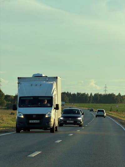 A white truck with a box trailer is traveling on a highway. Inside the truck, two men are visible in the cab. The truck is in the left lane of a two-lane road. To the right of the truck, a black sedan is traveling in the same direction. Further ahead, a white SUV is also on the highway. The road is paved with asphalt and has white lane markings. On the right side of the road, there are fields of grass and trees. Power lines and a transmission tower are visible in the background. The sky is clear with a pale blue hue. The scene appears to be set in a rural or semi-rural area, possibly Togashevo, Russia, given the context.