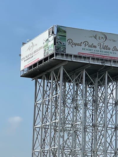 An elevated multi-panel billboard structure stands prominently against a clear blue sky in Igando Oloja, Nigeria. The structure consists of a massive grey metallic lattice framework of interconnected vertical and diagonal beams supporting the advertising panels. The primary billboard displays information for "ROYAL PALM VILLA IBEJU-LEKKI" and "RPV SPORTS CLUB," featuring images of various sports such as golf, tennis, and basketball, alongside the phrase "...satisfaction guaranteed." An adjacent billboard panel continues the "Royal Palm Villa Ibeju-Lekki RESORT AND RESIDENCES" advertisement, listing amenities like "GOLF • LAWN TENNIS • BASKETBALL • FOOTBALL • ICE SKATING," also displaying corresponding imagery. No individuals are present in the visible scene.