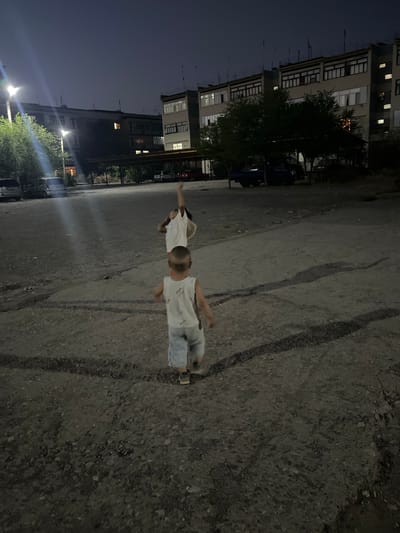 This photograph depicts a nighttime scene in a residential area in Şamaldı-Say, Kyrgyzstan. Two young children are visible in the foreground, one facing away from the camera, and another further in the background, with its arm raised. They are standing on a gravelly, uneven surface, likely a courtyard or parking area. In the background, apartment buildings are illuminated, suggesting it is evening or night. The sky is dark. The overall impression is of a quiet, possibly mundane, urban setting at night.