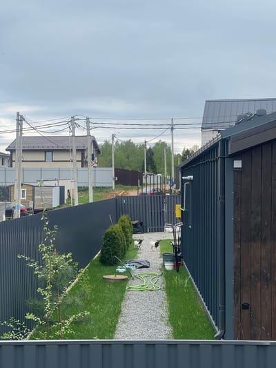 The image depicts a residential scene in Хлябово, Russia. The perspective is from an elevated position, looking across adjacent properties. In the foreground, a corrugated metal fence and the corner of a building with dark wood siding are visible. Between the fences, there is a narrow strip of land with gravel, grass, and various objects, including a green garden hose, lawn care equipment, and what appear to be plastic bottles and a trash bag. Two dogs, black and white, are present in the gravel area. Beyond the fences, there are more residential buildings, a gravel area, and an area of trees. Power lines stretch across the sky, supported by concrete poles. The sky is overcast and gray.