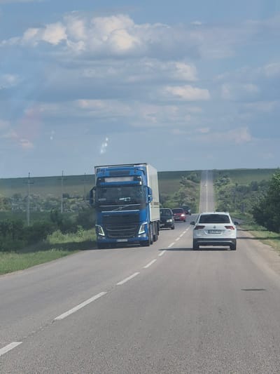 A blue Volvo semi-truck with a white trailer is traveling on a two-lane asphalt road, heading away from the camera. The truck is in the left lane. Ahead of the truck, in the right lane, is a white Volkswagen Tiguan. Further ahead in the distance, several other vehicles are visible, including a red car. The road is bordered by green grass and trees on both sides. In the background, rolling hills are covered with vegetation. The sky is blue with scattered white clouds. Electrical poles are visible along the side of the road. The scene appears to be a rural highway.