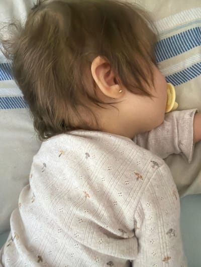 A close-up, top-down image captures a baby or young child sleeping peacefully on their side. The focus is on the child's head, with their face turned away, revealing their ear and fine, light brown hair. A small, golden stud earring is visible in the earlobe. Part of a yellow pacifier can be seen near the child's mouth.
The child is dressed in a pale-colored, likely cream or light pink, long-sleeved top or sleepsuit made of a soft, textured fabric. The garment features tiny patterned holes and small, scattered motifs in muted brown and grey tones, resembling leaves or insects.
The child rests on bedding consisting of a light-colored pillow or sheet with blue and white stripes, and another plain light-toned sheet or blanket. The scene conveys a sense of calm and rest, consistent with a child sleeping indoors, likely in a bedroom. The lighting is soft and diffused. No specific details within the image indicate the provided location of Bearpark, United Kingdom. There is no visible text.