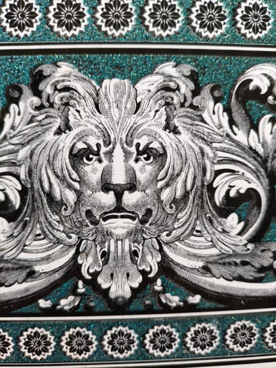 The image displays a close-up of a decorative item featuring a stylized lion's head in black and white. The lion's mane is elaborate and detailed, with swirling patterns that extend outwards. The lion's face is rendered with strong linework, emphasizing its features such as the eyes, nose, and mouth. Surrounding the lion's head are decorative scrolls and foliage, also in black and white.
The background of the decorative item is a textured teal-colored surface with visible glitter or small reflective particles, adding a shimmering effect. Below the central lion motif, there is a border with repeating black and white floral or star-like patterns, set against the same glittery teal background. This border is separated from the main image by a thin black and white striped band.
The overall style suggests a decorative plate, tile, or perhaps a panel from furniture or a box. The details are intricate, showcasing a classical or baroque aesthetic. There is no visible text in the image, and the lighting appears to be even, highlighting the texture and detail of the artwork. The scene is an artistic representation, not a photograph of a real-world event or setting.