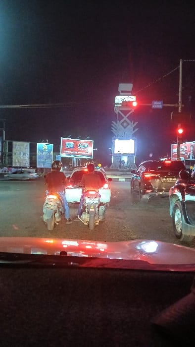 The image displays a nighttime urban street scene in Lamprehlam Teungoh, Indonesia, viewed from inside a stationary vehicle. The foreground shows the top of a dark dashboard with red light reflections. In the midground, traffic is halted, with a prominent red traffic light visible on the right. Two individuals, wearing dark clothing and helmets, are seated on separate motorbikes, positioned behind a white car. Their rear brake lights are illuminated. To the right of the white car, a dark colored SUV or truck is also stopped, displaying active taillights. In the background, multiple illuminated billboards are visible on both sides of the road, featuring text in an Indonesian context (e.g., "GRACIA," "HAPPY MOTY"). A large, illuminated, decorative monument or structure with a criss-cross pattern and a central screen stands in the center-right background, suggesting a roundabout or public square. The environment is dark, with primary illumination originating from vehicle lights, streetlights, and commercial signage.