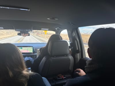 Inside a car, a view through the windshield shows a road stretching through a landscape of brown fields under a partially cloudy sky. The rearview mirror reflects the driver's face. Two people are visible in the back seats, one with long brown hair and another with short dark hair wearing a scarf. A navigation screen is visible on the dashboard. The interior of the car is gray.