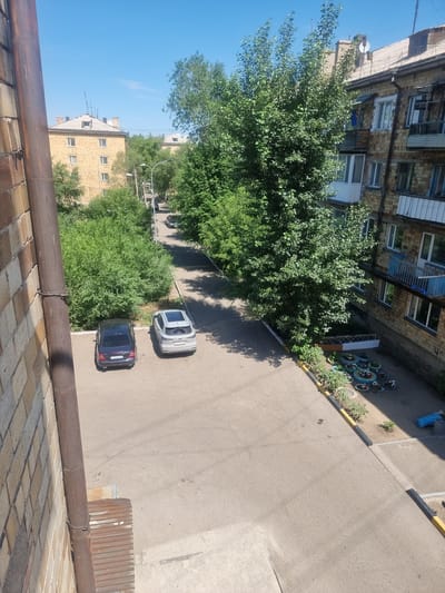 The image depicts a sunny day in a residential area. Two cars are parked on an asphalt surface in front of a multi-story brick building. One car is dark blue, and the other is silver with a sunroof. Adjacent to the parking area is a patch of greenery with several young plants and a collection of flower pots made from repurposed tires. Beyond this, more trees and bushes line the street. In the background, another building, also brick, can be seen, with trees in front of it. The sky is clear blue. The image appears to be taken from an elevated position, possibly a balcony or window. The scene is set in the city of Saran, Kazakhstan, likely within the administrative area of the city administration, given the urban residential setting. There are no people visible in the image.